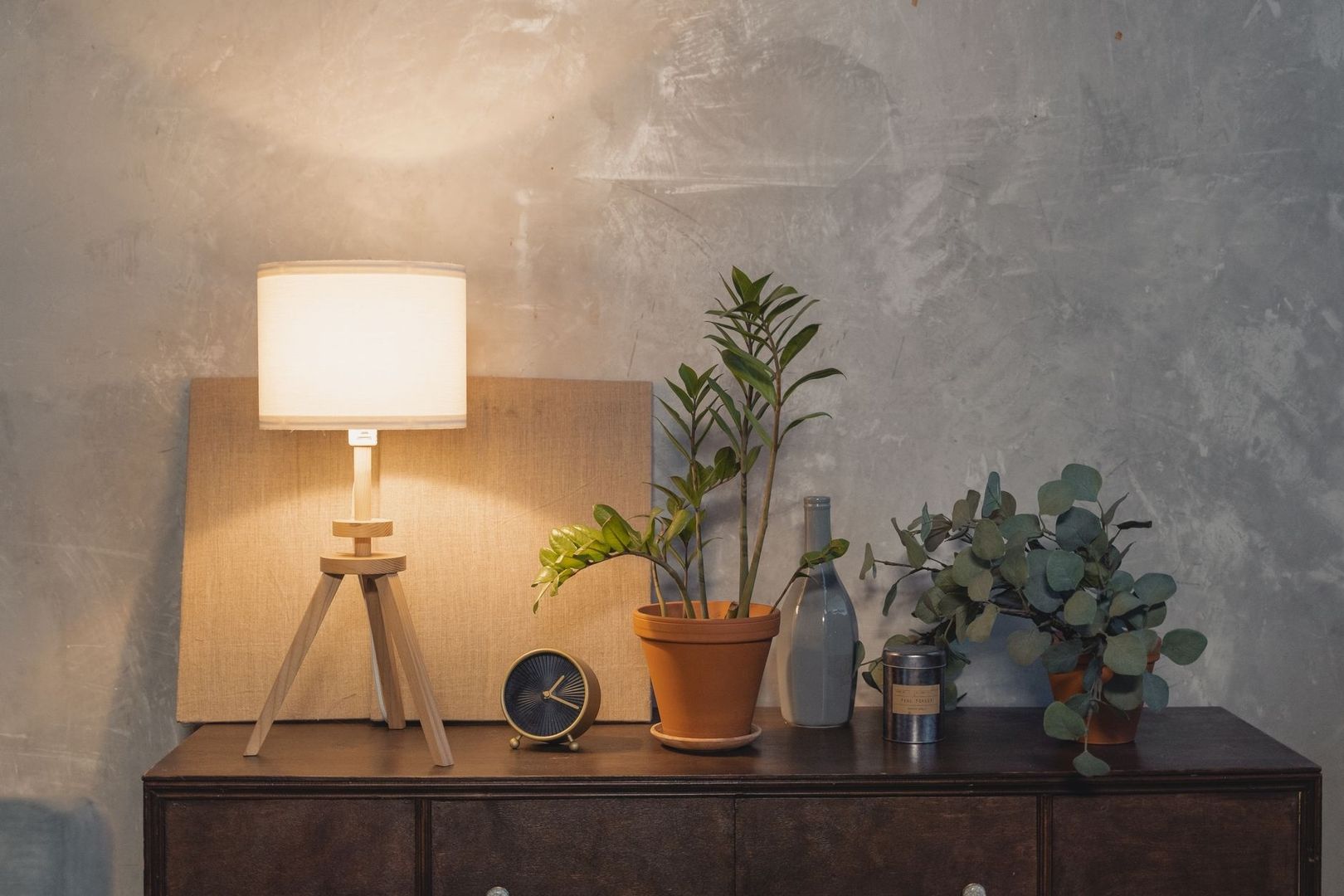 Varm bordslampa, växter och klocka på mörk träbyrå